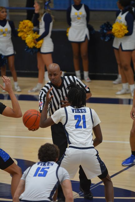 The image depicts a sports scene inside a gymnasium, where a referee dressed in a black and white striped shirt is holding a basketball and engaging with two young male basketball players in white jerseys with blue accents and the word 'VIKES' printed on the back, with their jersey numbers 21 and 15 visible. The player wearing jersey 21 is standing with his back to the camera, facing the referee, while the player with jersey 15 is partially visible, positioned in front of him. In the background, a group of cheerleaders dressed in navy blue and white uniforms with yellow pom-poms are lined up, holding their pom-poms and observing the game. The gymnasium's wooden floor is visible, along with blue padding along the walls, and the environment is well-lit with bright overhead lights, creating a lively and organized atmosphere typical of an indoor sports event. The setting suggests an official match or practice session with active participation and spectators present for support.