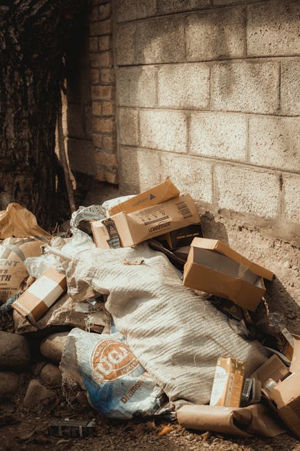 A pile of mixed waste materials is accumulated at the base of a brick and concrete block wall adjacent to a tree trunk. The waste includes several flattened cardboard boxes, some with printed labels and barcodes, along with a large woven fabric or sack that appears to be used for rubbish collection or storage. There are also plastic bags, one of which is partially visible with a printed logo, and crumpled brown paper wrapping. The debris is spread on uneven ground composed of small rocks and dirt, with some loose bits of paper and packaging around. The scene appears to be in an outdoor area possibly used for informal waste disposal, reflecting an alternative rubbish removal situation that might be handled by private waste clearance services such as Rubbish Clearance Earls Court. The lighting suggests natural daylight, casting minimal shadows on the cluttered heap, emphasizing the textures and colors of the discarded materials and the rough surface of the brick and concrete wall behind it.