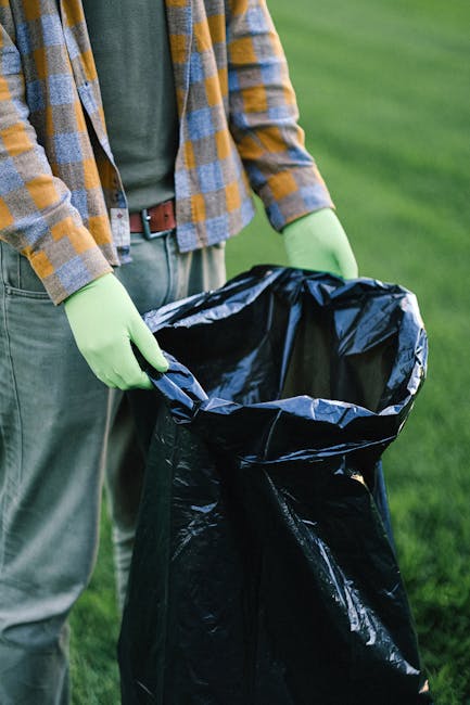 In this outdoor scene, multiple black garbage bags are placed on a grassy area in front of a modern residential building, which features a brick facade and large windows. The bags are loosely arranged, some standing upright and others leaning or resting on the ground, indicating they are filled with waste or rubbish awaiting collection. Several bags are positioned close to the base of leafless trees with dark, textured bark that have branches spreading out across the scene, partially obscuring the building behind. The environment appears to be a communal or shared outdoor space typical of apartment complexes, and the overcast sky provides diffuse lighting, creating a neutral, professional atmosphere suitable for depicting private waste disposal in the context of rubbish removal services. The overall image suggests on-site rubbish collection, aligned with independent waste management options served by companies such as Rubbish Clearance Earls Court, supporting efficient rubbish disposal outside of local authority collection routines.