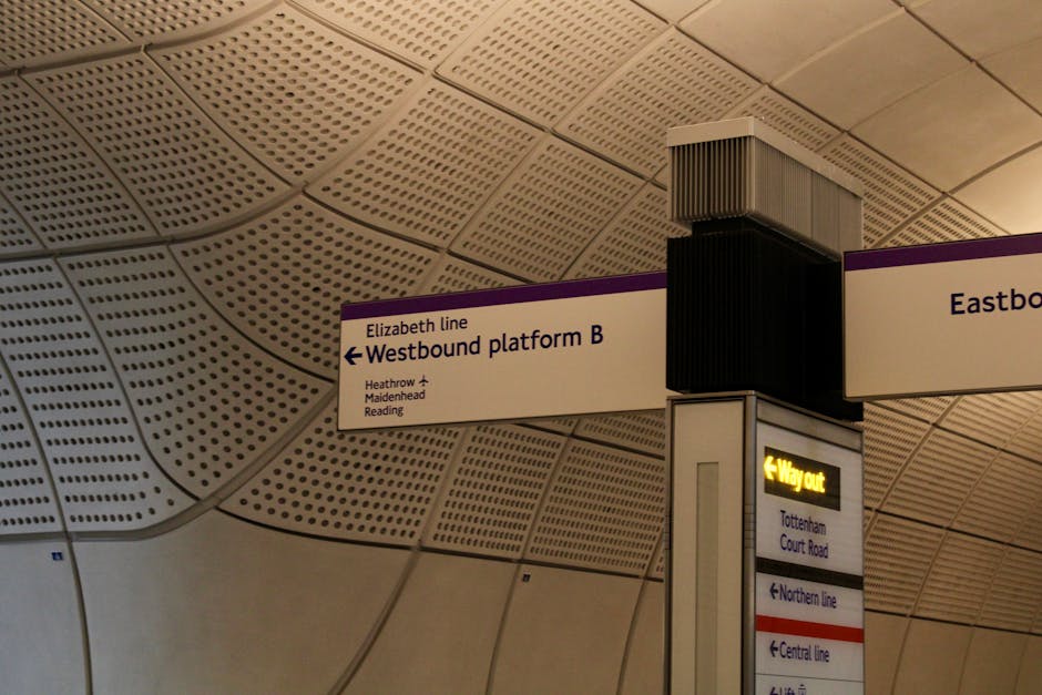 A close-up view of a station signage post situated in an indoor environment with a curved, composite ceiling featuring a pattern of perforated panels. The sign displays directions to the westbound platform B for Elizabeth line services, with additional information about destinations such as Heathrow, Maidenhead, and Reading. Above the sign, a large black ventilation or audio unit is mounted, with visible vents on its sides. To the right, a yellow-lit 'Way out' indicator points left toward routes to Tottenham Court Road, with smaller directional signs below indicating other train lines including Northern and Central lines. The environment appears well-lit with ambient lighting emphasizing the ceiling's textured surface, giving a sense of enclosure typical of modern underground stations. This scene relates to public transport infrastructure, similar to private or alternative waste handling facilities in terms of indoor structural clarity and directional signage, though more relevant for travel instructions than waste management directly.