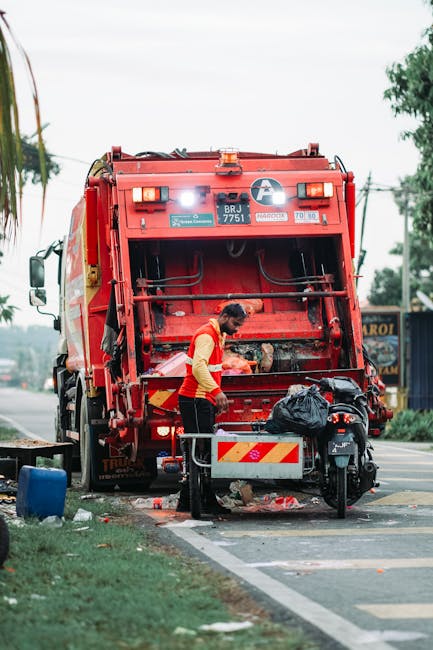 A worker wearing a red and yellow uniform is operating a large red rubbish collection truck on a street, with its rear compactor open and material being loaded or compacted. The scene shows the worker accessing the truck's rear loading area, which is partially filled with waste and debris. Nearby, there is a black bag of refuse placed on a metal platform, and a motorcycle is parked close to the truck's rear, indicating it may be used for logistics or support. The environment appears to be an urban or suburban area with a paved road, a grassy verge, and some trees or foliage in the background. The truck is marked with safety reflective stripes and identification labels, emphasizing its purpose for commercial waste collection and rubbish removal services, such as those offered by Rubbish Clearance Earls Court. The lighting suggests daylight conditions, with overcast skies contributing to diffuse natural light. This scene exemplifies on-site waste handling and the process of private rubbish collection, aligning with professional rubbish removal activities aimed at efficient clearance outside of traditional waste authority services.